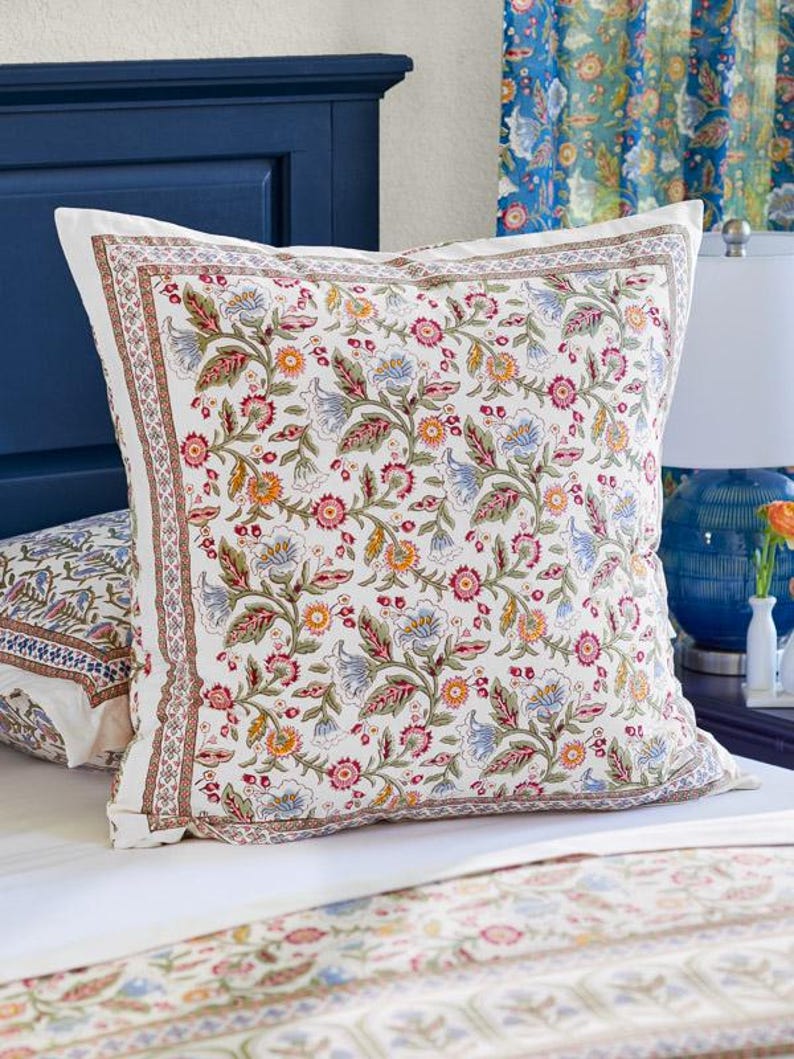 May include: A square pillow with a white background and a colorful floral pattern. The pattern includes red, blue, yellow, and green flowers and leaves. The pillow has a decorative border and is placed on a bed with a matching floral patterned bedspread.