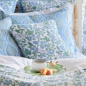 May include: A bed with blue and white floral patterned bedding, including pillows and a duvet. A white mug, pastry, and decorative bird are on a green leaf-shaped tray. The headboard is made of woven wicker.
