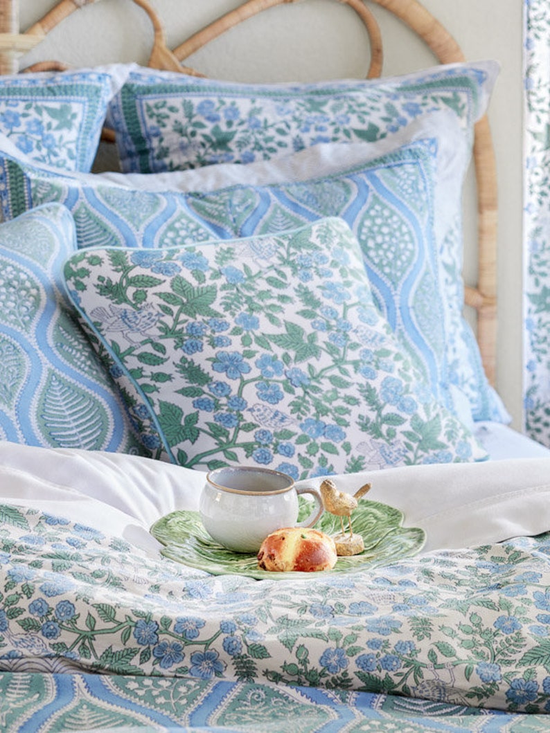 May include: A bed with blue and white floral patterned bedding, including pillows and a duvet. A white mug, pastry, and decorative bird are on a green leaf-shaped tray. The headboard is made of woven wicker.