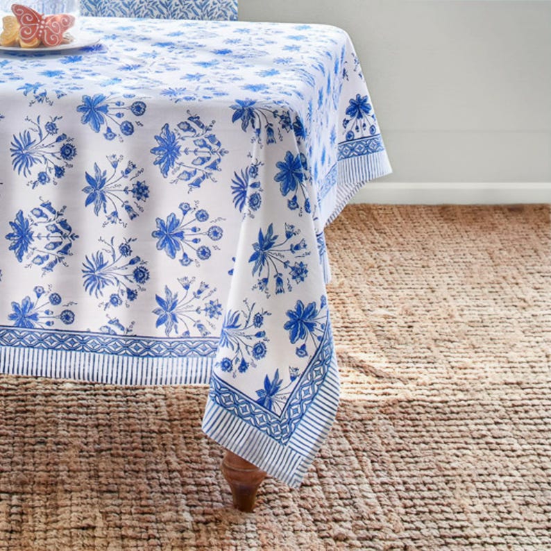 May include: White tablecloth with a blue floral pattern. The tablecloth has a repeating design of blue flowers and leaves. The border features a striped and geometric pattern. The tablecloth is draped over a wooden table.