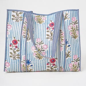 May include: A rectangular tote bag featuring a white background with vertical blue stripes and a repeating floral pattern in shades of pink, red, and green. The bag includes two matching fabric handles with blue and white striped trim.