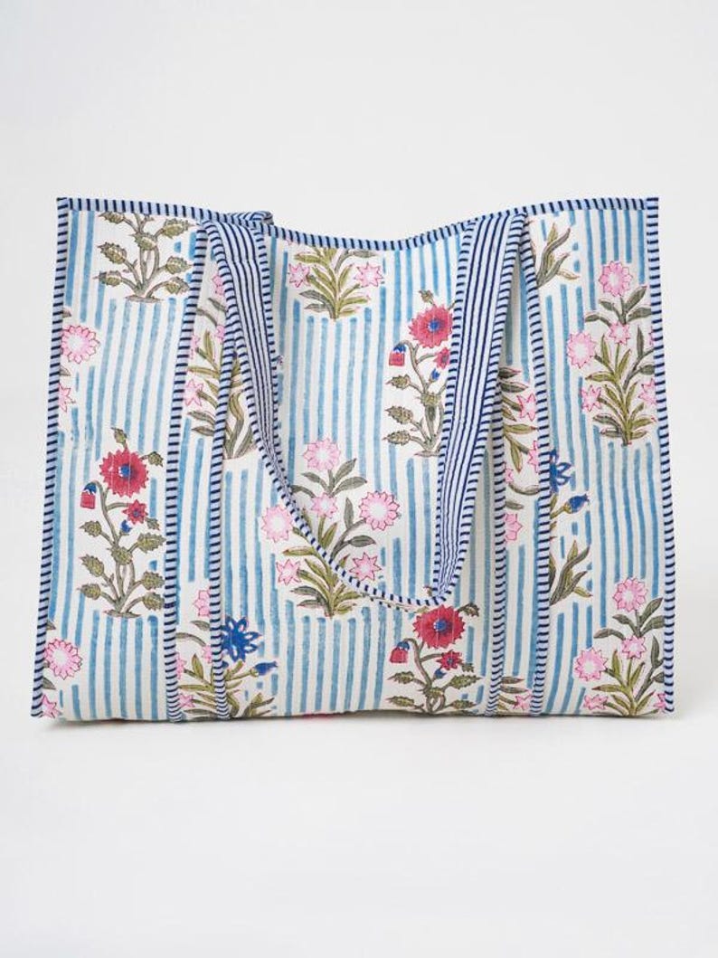 May include: A rectangular tote bag featuring a white background with vertical blue stripes and a repeating floral pattern in shades of pink, red, and green. The bag includes two matching fabric handles with blue and white striped trim.