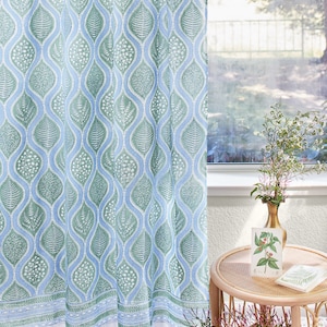Op de afbeelding: Een transparant blauw en groen gordijn met een bladmotief. Het gordijn hangt in een raam met uitzicht op een tuin.