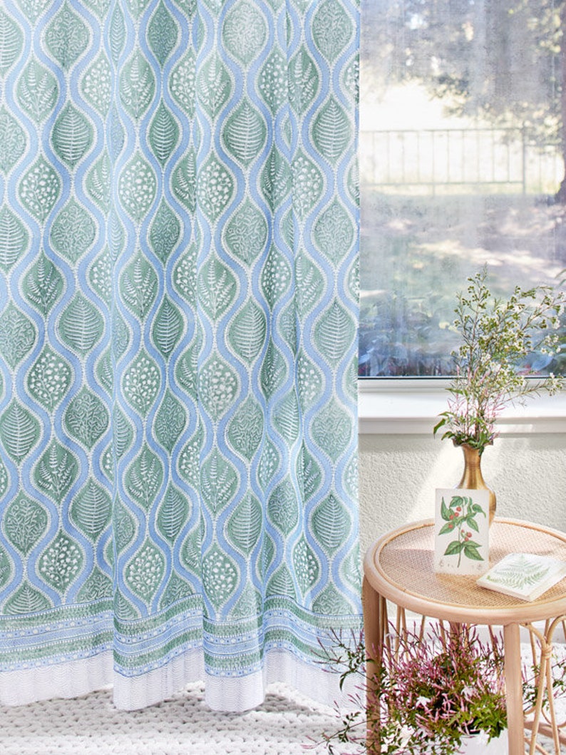 Op de afbeelding: Een transparant blauw en groen gordijn met een bladmotief. Het gordijn hangt in een raam met uitzicht op een tuin.
