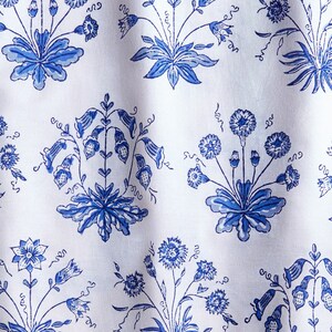May include: White fabric with a repeating pattern of blue floral bouquets. The bouquets feature various flower types and leaf designs. The fabric has a slightly wrinkled texture, suggesting it is made of a soft material like cotton or linen.