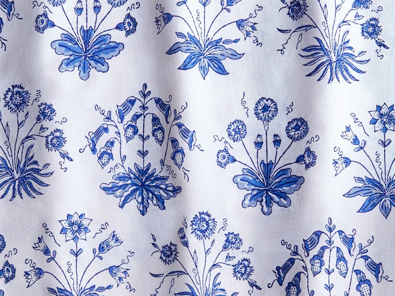 May include: White fabric with a repeating pattern of blue floral bouquets. The bouquets feature various flower types and leaf designs. The fabric has a slightly wrinkled texture, suggesting it is made of a soft material like cotton or linen.