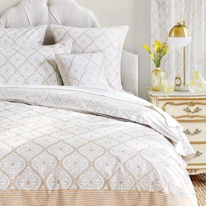 May include: A white and beige patterned duvet cover with a tan and white striped border. The duvet cover is on a bed with a white headboard and two patterned pillows.