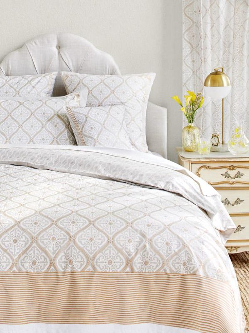 May include: A white and beige patterned duvet cover with a tan and white striped border. The duvet cover is on a bed with a white headboard and two patterned pillows.