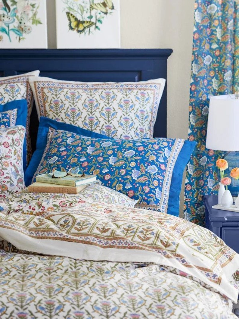 May include: A bed with a navy blue headboard and bedding featuring floral patterns in blue, white, and pink. The pillows and duvet cover display intricate designs. Two framed paintings with floral and butterfly motifs hang above the bed.