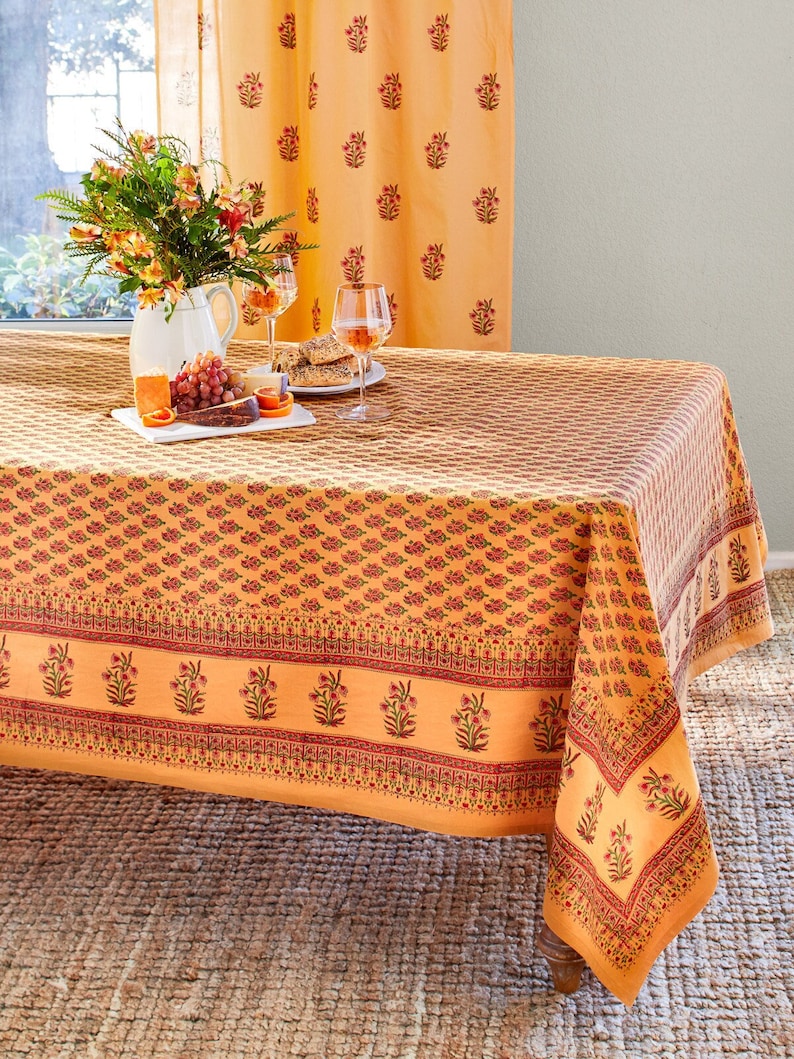 May include: A yellow and red floral tablecloth with a border of smaller floral designs. The tablecloth is laid on a table with a vase of flowers, a plate of fruit, and two glasses of wine.