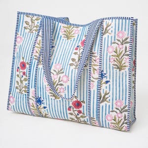 May include: A rectangular tote bag featuring a white background with vertical blue stripes and a floral pattern in shades of pink, red, and green. The bag includes two matching striped fabric handles and blue and white striped trim along the edges.