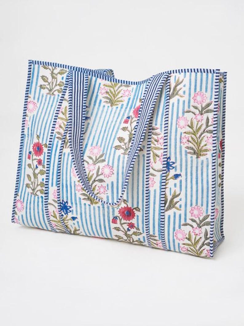 May include: A rectangular tote bag featuring a white background with vertical blue stripes and a floral pattern in shades of pink, red, and green. The bag includes two matching striped fabric handles and blue and white striped trim along the edges.