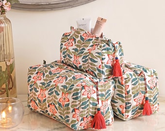 Puede incluir: Tres estuches de tela acolchada con fondo blanco y un patrón repetitivo de flores rosa coral y hojas verde oscuro. Cada estuche rectangular incluye un tirador de cremallera con borla roja y contiene tubos cosméticos.
