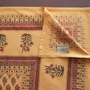 May include: A yellow cotton tablecloth with a red, green, and white floral pattern. The tablecloth has a border with a repeating design. The tablecloth is folded in half and has a tag that says "Handloom Cotton Tablecloth"