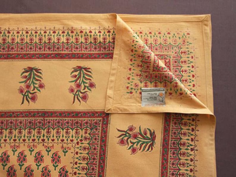 May include: A yellow cotton tablecloth with a red, green, and white floral pattern. The tablecloth has a border with a repeating design. The tablecloth is folded in half and has a tag that says "Handloom Cotton Tablecloth"