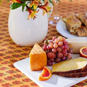 May include: A white platter with a variety of cheeses, grapes, and blood orange slices. The platter is on a table with a floral pattern tablecloth.