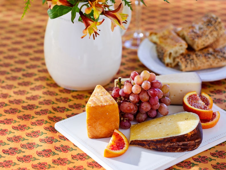 May include: A white platter with a variety of cheeses, grapes, and blood orange slices. The platter is on a table with a floral pattern tablecloth.
