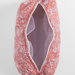 May include: A quilted cosmetic bag in a dusty rose color featuring a white floral pattern. The bag has a white zipper closure, an open interior with white lining, and a small decorative tassel attached to the zipper pull at the top end.