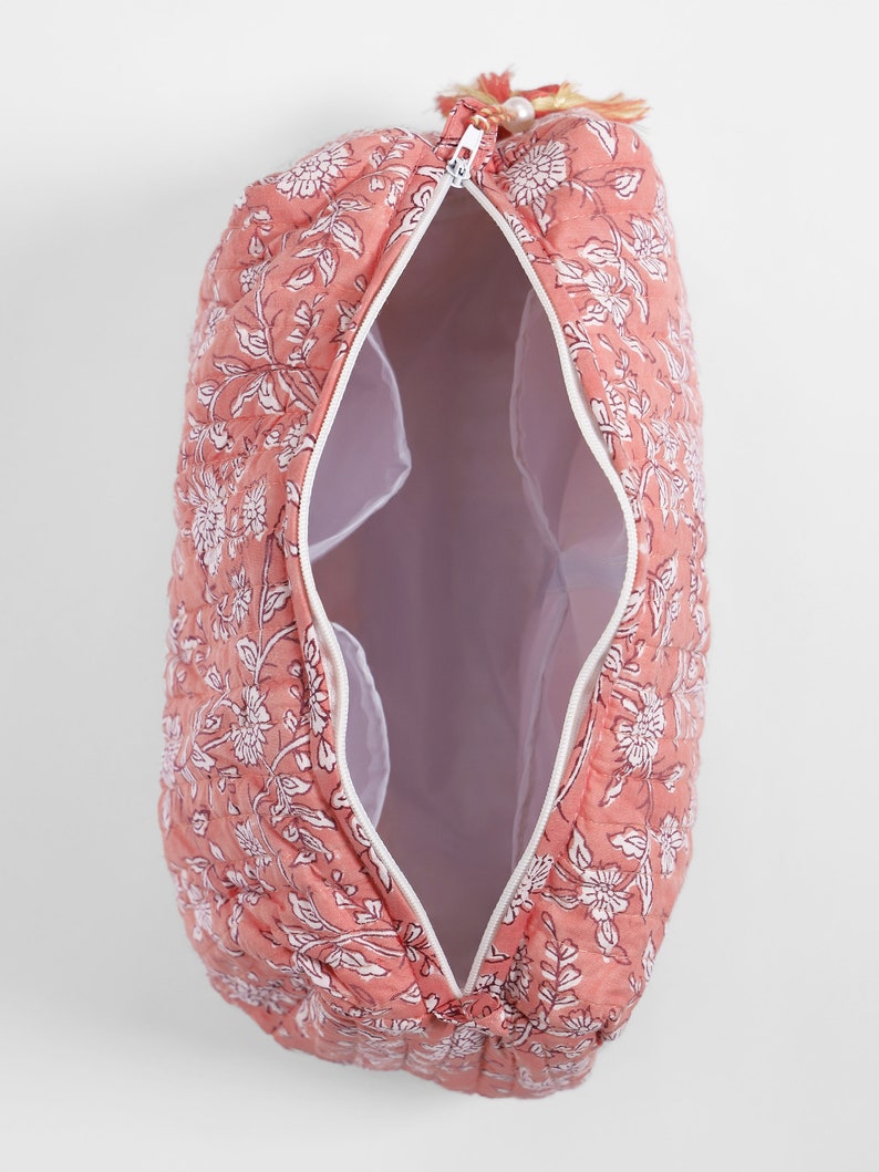 May include: A quilted cosmetic bag in a dusty rose color featuring a white floral pattern. The bag has a white zipper closure, an open interior with white lining, and a small decorative tassel attached to the zipper pull at the top end.