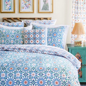 May include: A white bed with a blue and orange patterned duvet cover. The duvet cover has a geometric pattern with a repeating design of stars and flowers. The bed is made up with white pillows and a blue and orange patterned throw pillow. There are three framed pictures on the wall behind the bed.