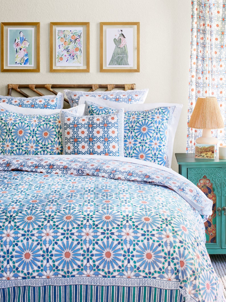 May include: A white bed with a blue and orange patterned duvet cover. The duvet cover has a geometric pattern with a repeating design of stars and flowers. The bed is made up with white pillows and a blue and orange patterned throw pillow. There are three framed pictures on the wall behind the bed.