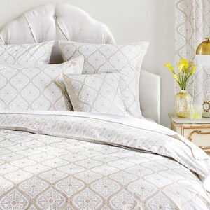 May include: A white bed with a patterned duvet cover and matching pillowcases. The bed has a headboard with a tufted design. There is a nightstand with a lamp and flowers next to the bed.
