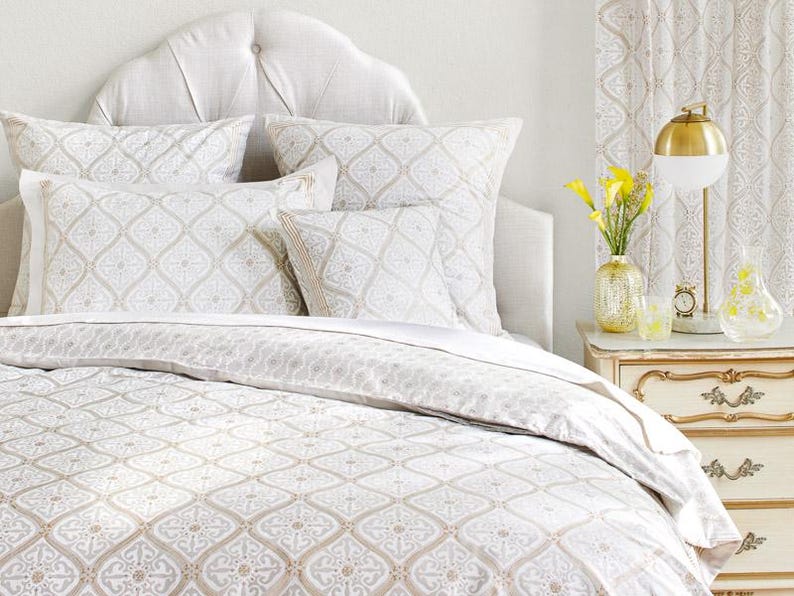 May include: A white bed with a patterned duvet cover and matching pillowcases. The bed has a headboard with a tufted design. There is a nightstand with a lamp and flowers next to the bed.