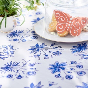 May include: A white tablecloth with a blue floral pattern is the backdrop for a plate of butterfly-shaped cookies under a glass cloche. A small potted plant sits to the left. The cookies are decorated with white icing.