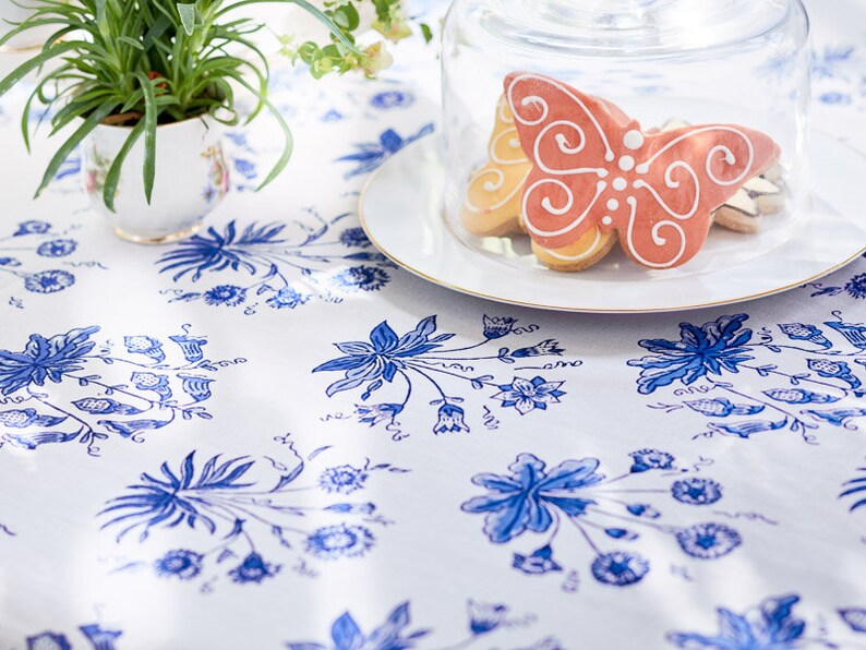 May include: A white tablecloth with a blue floral pattern is the backdrop for a plate of butterfly-shaped cookies under a glass cloche. A small potted plant sits to the left. The cookies are decorated with white icing.