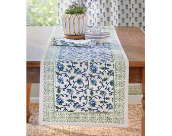 May include: A white table runner with a blue floral pattern and green border. The runner is placed on a wooden table, with matching napkins and a vase of flowers. The floral design includes blue flowers and green leaves.