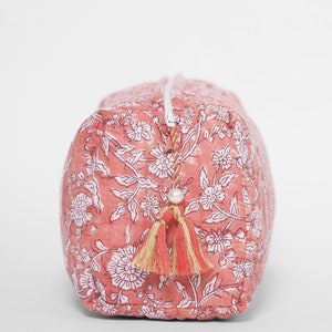 May include: A rectangular quilted pouch in a dusty rose pink color featuring a white floral block print pattern. The pouch has a white zipper closure accented with a decorative pull featuring a small white bead and two dual-toned fabric tassels.