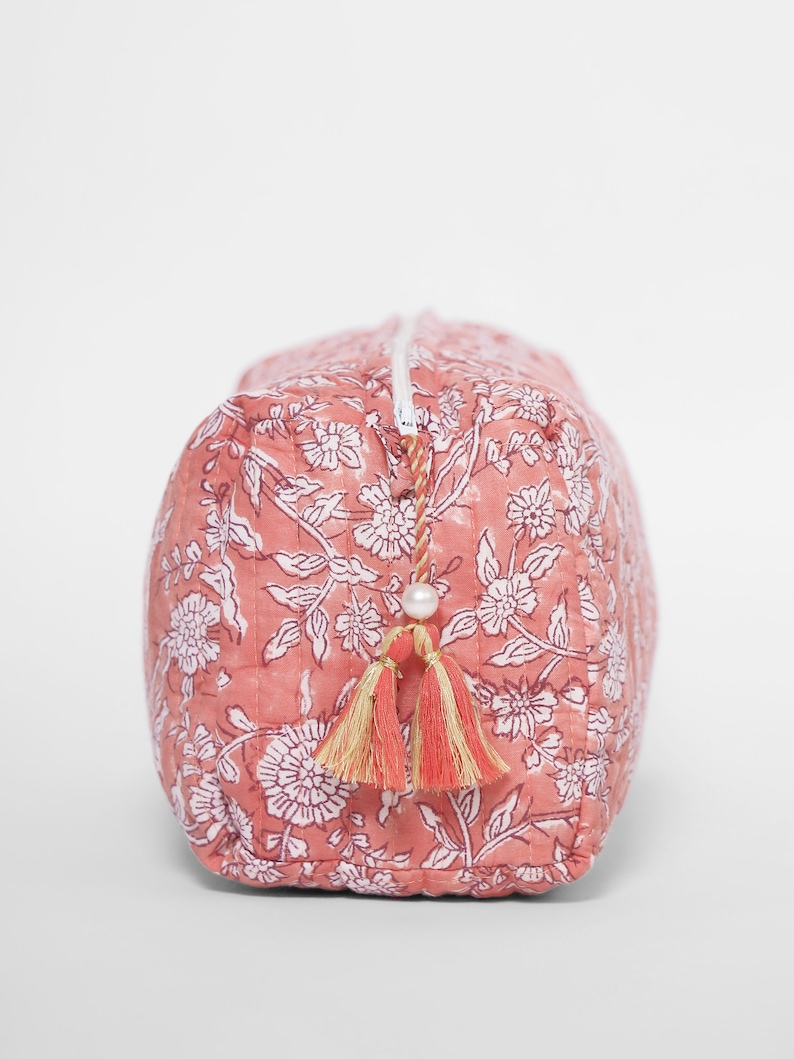 May include: A rectangular quilted pouch in a dusty rose pink color featuring a white floral block print pattern. The pouch has a white zipper closure accented with a decorative pull featuring a small white bead and two dual-toned fabric tassels.