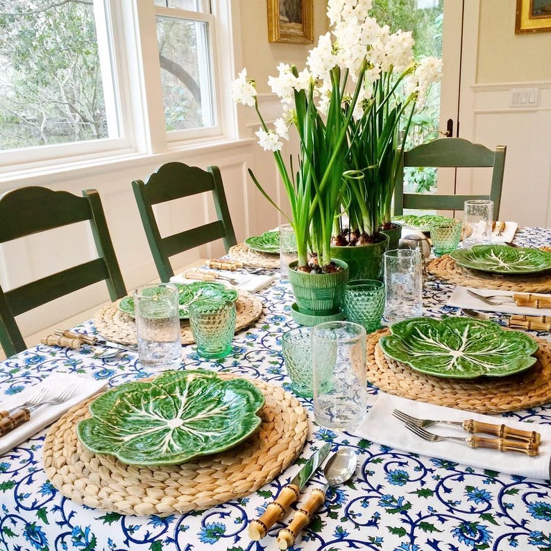K&ouml;nnte beinhalten: Eine Tischdekoration mit einer wei&szlig;en Tischdecke mit blauem Blumenmuster. Gr&uuml;ne, kohlf&ouml;rmige Teller sind auf geflochtenen Platzsets platziert. Gr&uuml;ne Glasbecher und Bambusbesteck stehen auf dem Tisch. Ein Strau&szlig; wei&szlig;er Narzissen in einem gr&uuml;nen Topf steht in der Mitte des Tisches.