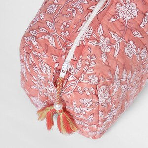 May include: A quilted rectangular pouch in a dusty rose pink color featuring a white floral block print pattern. The pouch includes a white zipper closure with a decorative pull featuring a small white bead and two-toned pink and yellow tassels.