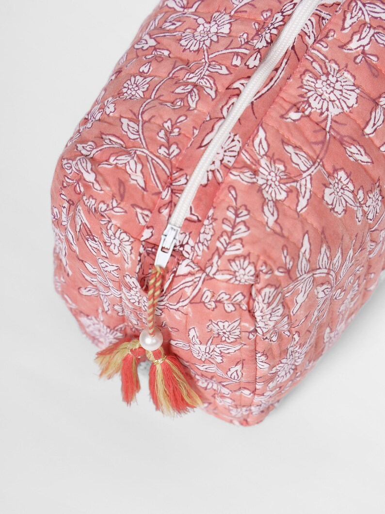 May include: A quilted rectangular pouch in a dusty rose pink color featuring a white floral block print pattern. The pouch includes a white zipper closure with a decorative pull featuring a small white bead and two-toned pink and yellow tassels.
