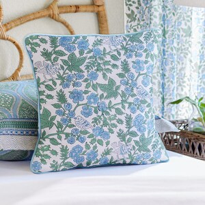 May include: Square throw pillow with a floral pattern in blue and green on a white background. The pillow has a light blue border and features a design of flowers, leaves, and birds. The pillow is on a white bed.