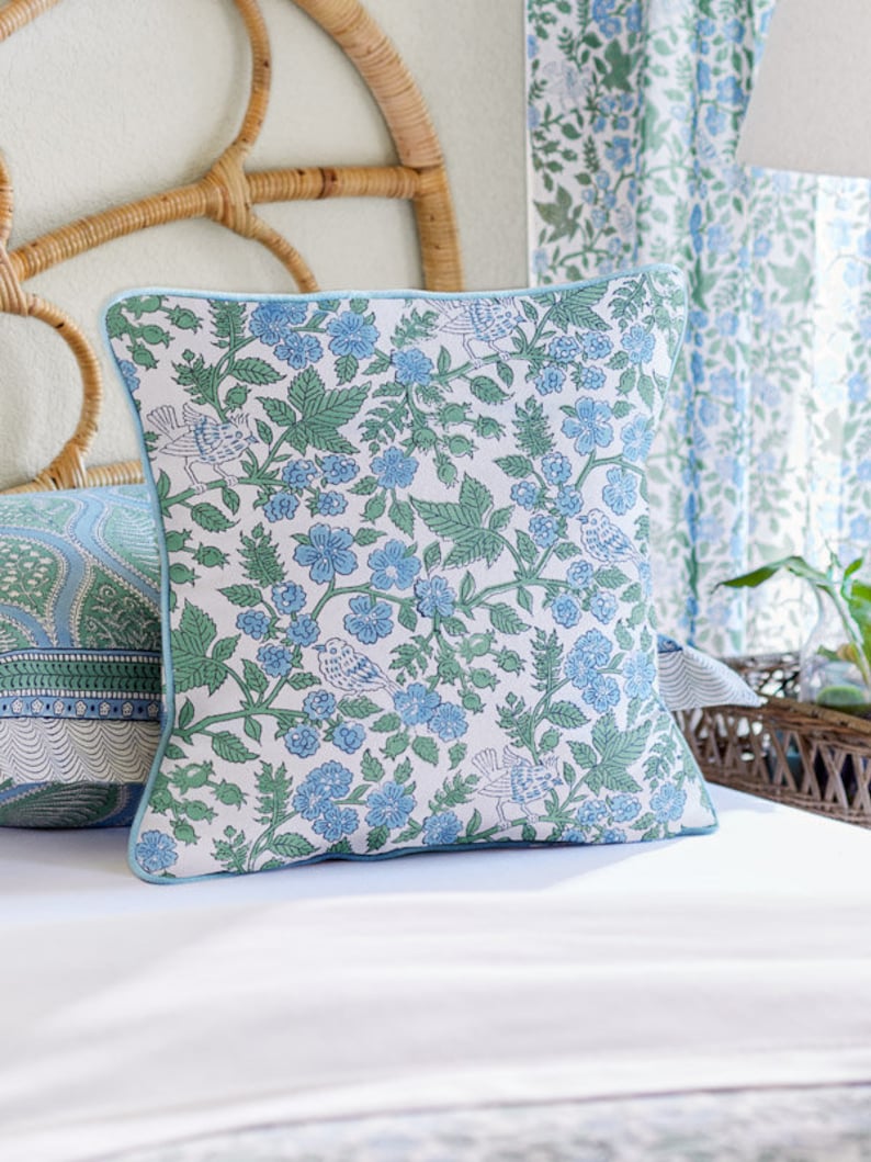 May include: Square throw pillow with a floral pattern in blue and green on a white background. The pillow has a light blue border and features a design of flowers, leaves, and birds. The pillow is on a white bed.