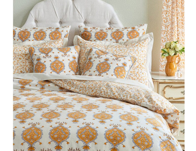 May include: A white bed with a white headboard and a white and yellow patterned duvet cover. The duvet cover has a paisley design with yellow and blue accents. There are several pillows on the bed, all with the same paisley design.