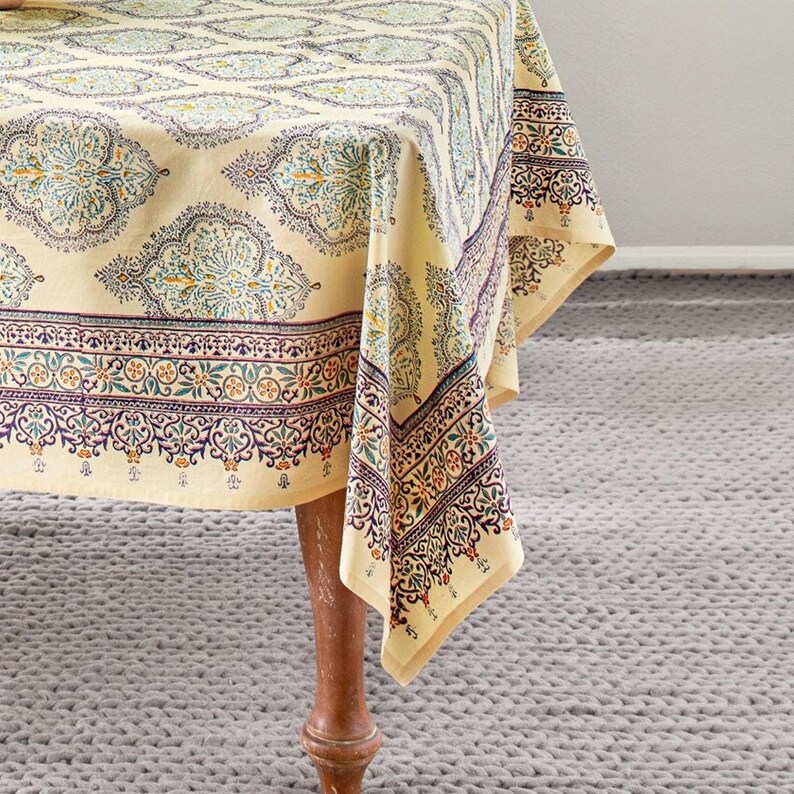 Puede incluir: Un mantel beige con un patr&oacute;n detallado y repetitivo en verde azulado, morado y naranja. El mantel est&aacute; drapeado sobre una mesa de madera, con la esquina de la tela colgando. El patr&oacute;n incluye elementos florales y geom&eacute;tricos.