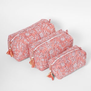 May include: A set of three rectangular quilted pouches in varying sizes, featuring a coral pink base with a white floral and vine print. Each pouch has a white zipper closure accented with a decorative tassel featuring a small white bead.