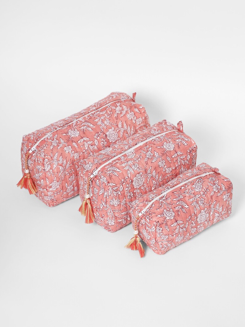 May include: A set of three rectangular quilted pouches in varying sizes, featuring a coral pink base with a white floral and vine print. Each pouch has a white zipper closure accented with a decorative tassel featuring a small white bead.