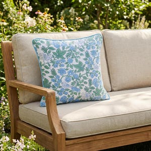 May include: A square throw pillow with a floral pattern in shades of blue and green, set against a cream background. The pillow has a light blue border and is placed on a wooden outdoor sofa with cream-colored cushions. The sofa is surrounded by greenery.