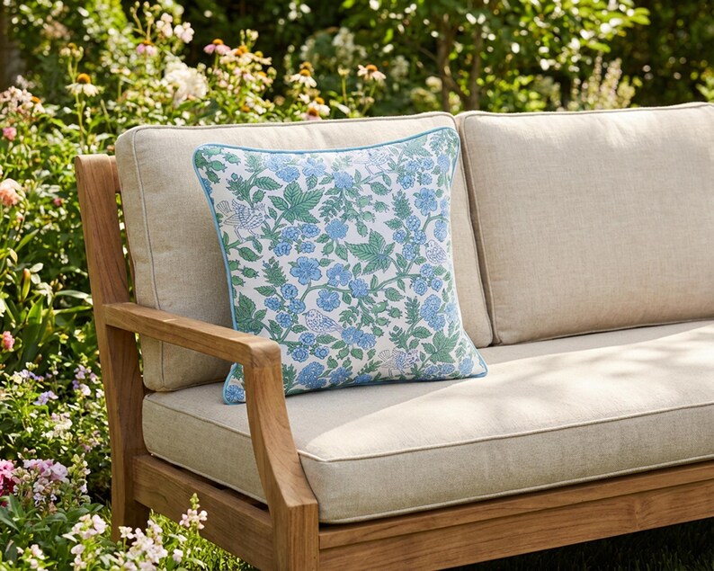 May include: A square throw pillow with a floral pattern in shades of blue and green, set against a cream background. The pillow has a light blue border and is placed on a wooden outdoor sofa with cream-colored cushions. The sofa is surrounded by greenery.