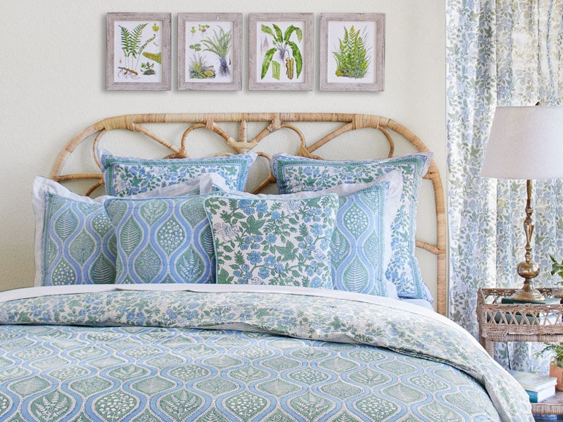 May include: A bedroom scene featuring a bed with a woven headboard, adorned with blue and green patterned bedding and pillows. Above the bed, four framed botanical prints hang on the wall. Matching curtains and a bedside lamp complete the coordinated decor.