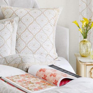 May include: A white bed with a patterned pillow and a book with a floral design on top of the bedding. A vase with yellow flowers sits on a nightstand next to the bed.