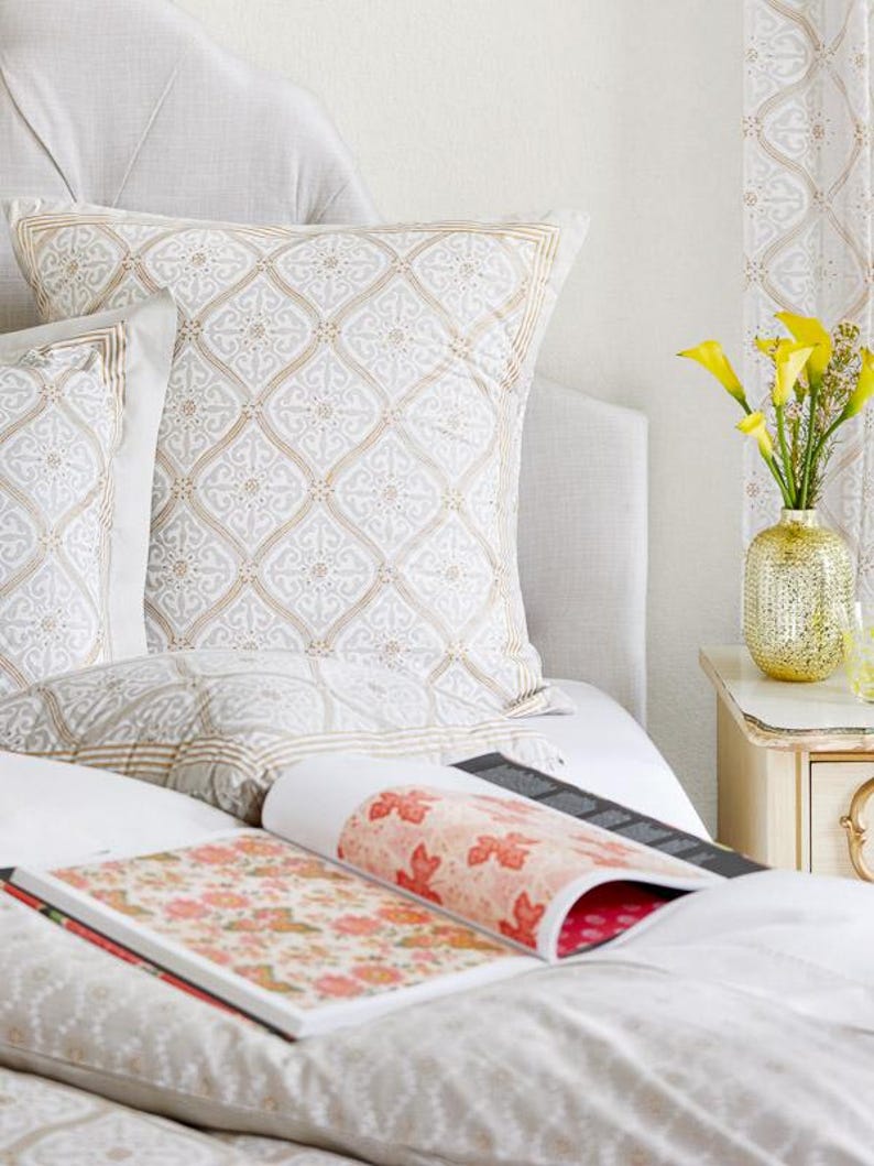 May include: A white bed with a patterned pillow and a book with a floral design on top of the bedding. A vase with yellow flowers sits on a nightstand next to the bed.