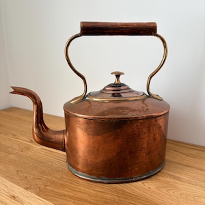Könnte beinhalten: Ein vintage Teekessel aus Kupfer mit zylindrischem Körper, gebogenem Ausguss und einem Messinggriff mit Holzgriffstück. Der Kessel verfügt über einen abnehmbaren Deckel mit einem kleinen Messingknauf. Die Metalloberfläche zeigt eine Patina.