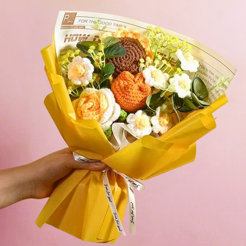 May include: A yellow paper bouquet of crocheted flowers. The bouquet includes orange, brown, and white flowers. The flowers are wrapped in yellow paper with a white ribbon.
