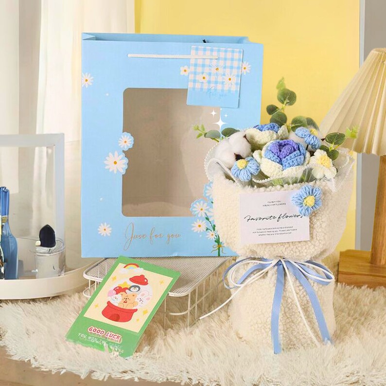 May include: A blue and white gift bag with a crocheted flower bouquet inside. The bouquet is wrapped in white faux fur and tied with a blue ribbon. The gift bag has a window that shows the bouquet inside. The gift bag has the text "Just for you" printed on it. The bouquet has a card that says "Favorite flowers".
