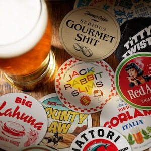 May include: A collection of colorful round coasters with various designs and text. Some of the coasters feature humorous phrases like "Serious Gourmet Shit" and "Titty Twister". Other coasters feature images of a woman smoking a cigarette, a burger, and a cartoon rabbit.